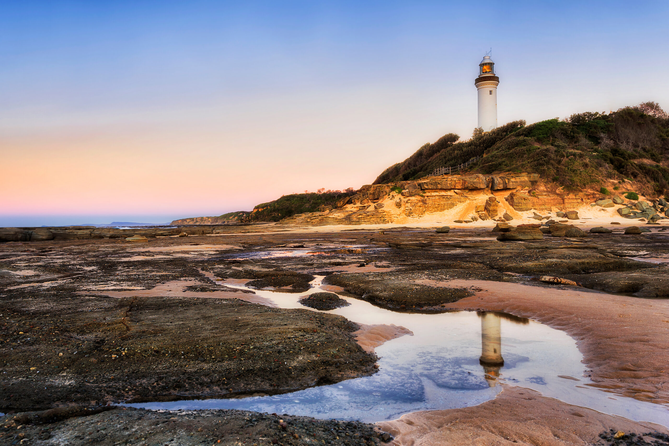 Seabed at low tide leaving salt water puddle to reflect white lighthouse and pink sky at sunrise on Australian Pacific coast - Norah headland, the Central Coast.